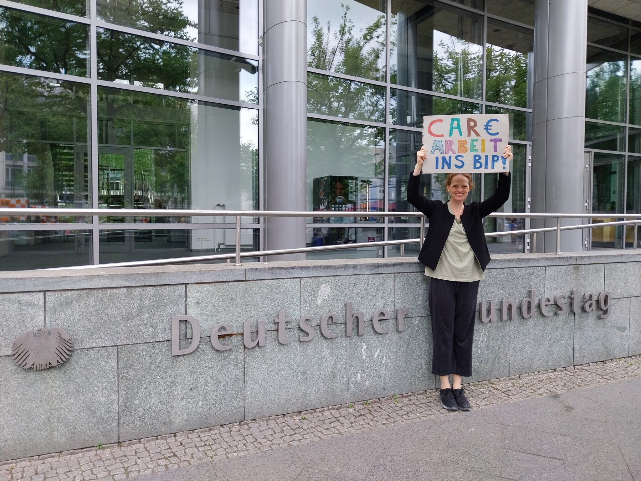 Hanna Dude am Seiteneingang des Deutschen Bundestages in Berlin, Hanna hält Plakat mit Text 'Care Arbeit ins BIP' hoch